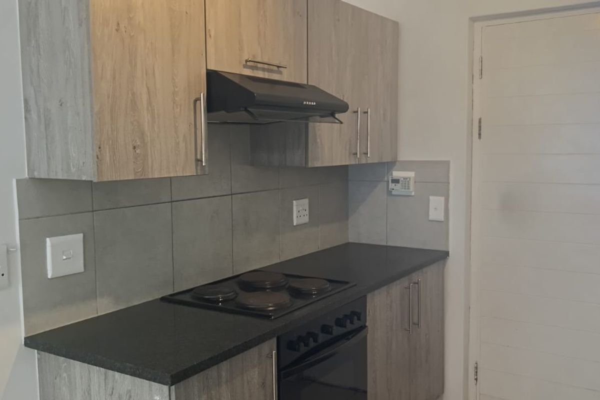 A clean, modern kitchen featuring wood-look cabinetry, dark countertops, a built-in electric hob, oven, and extractor fan with a neutral tiled backsplash.