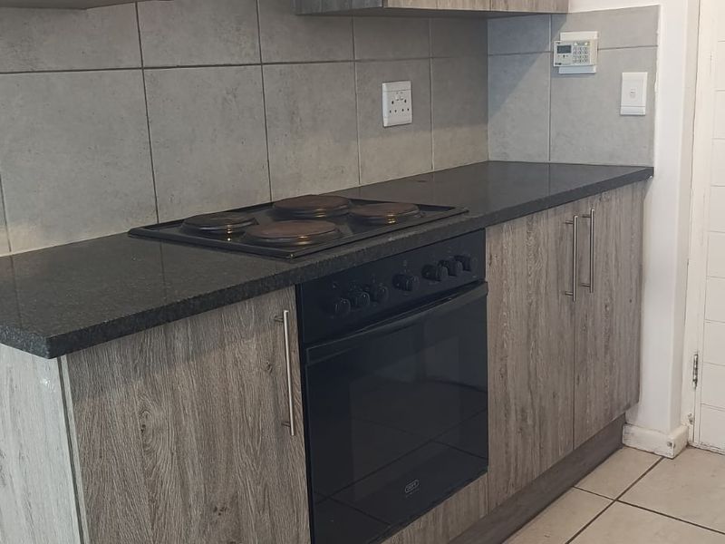 A functional kitchen area featuring modern grey laminate cabinetry, a dark stone countertop, built-in oven, electric hob, and neutral tiled backsplash.
