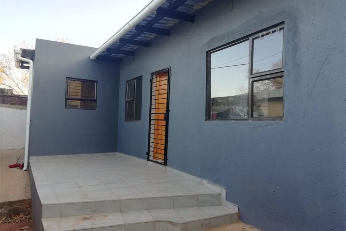 A rear exterior view featuring a tiled patio area, dark grey painted walls, and aluminium window frames with security bars.