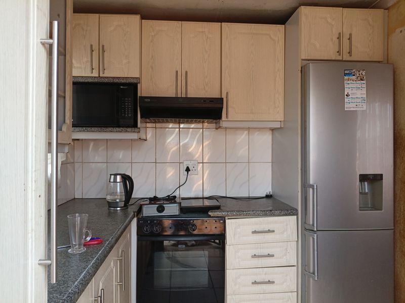 A compact kitchen featuring light wood-tone cabinetry, tiled backsplash, and integrated appliances, showing signs of wear and requiring cosmetic updates.