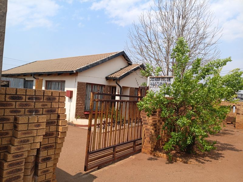 Front exterior view of a single-story brick house featuring a metal driveway gate and a tiled roof, with a prominent business sign on-site.