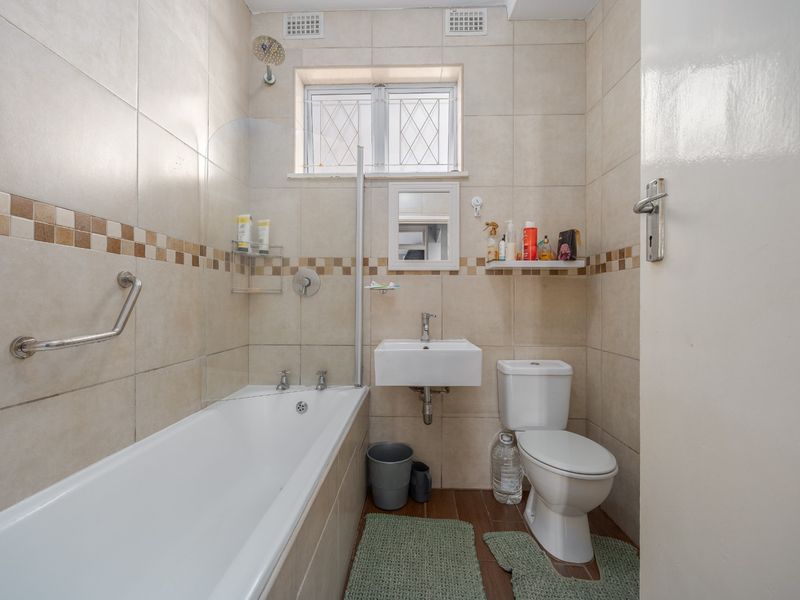 Compact bathroom featuring a bathtub with shower screen, wall-mounted basin, and toilet, finished with neutral wall tiling.