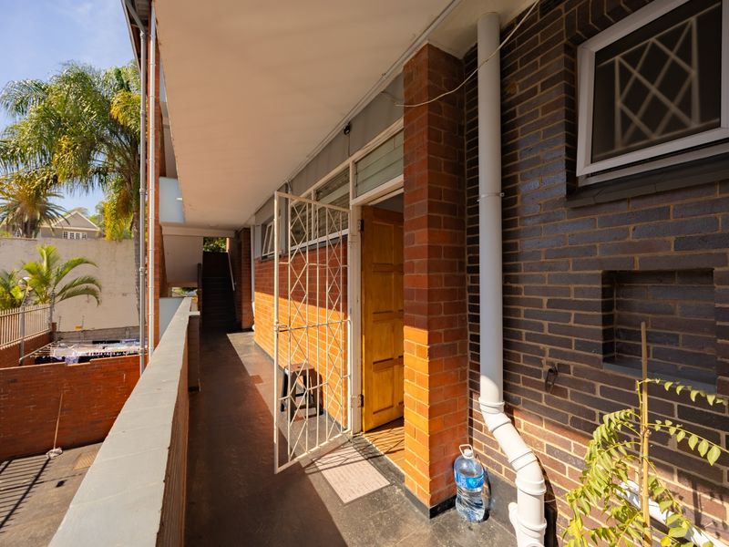 An exterior balcony walkway featuring brick masonry, a security gate, and a functional downpipe system overlooking the apartment complex grounds.
