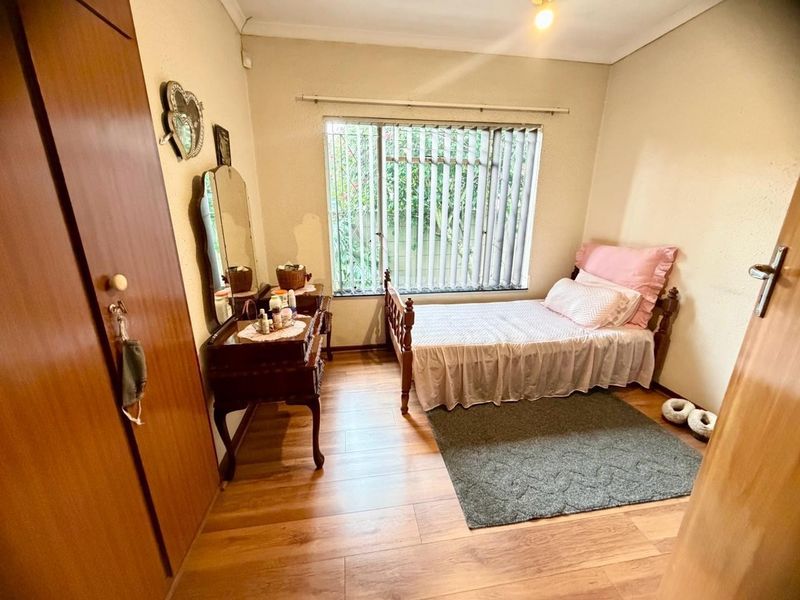 A functional bedroom featuring laminate flooring, a built-in wardrobe, and a window fitted with security bars and vertical blinds.