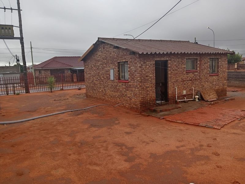 A detached face brick house featuring a corrugated metal roof and basic external infrastructure, situated on an unpaved plot with perimeter fencing.