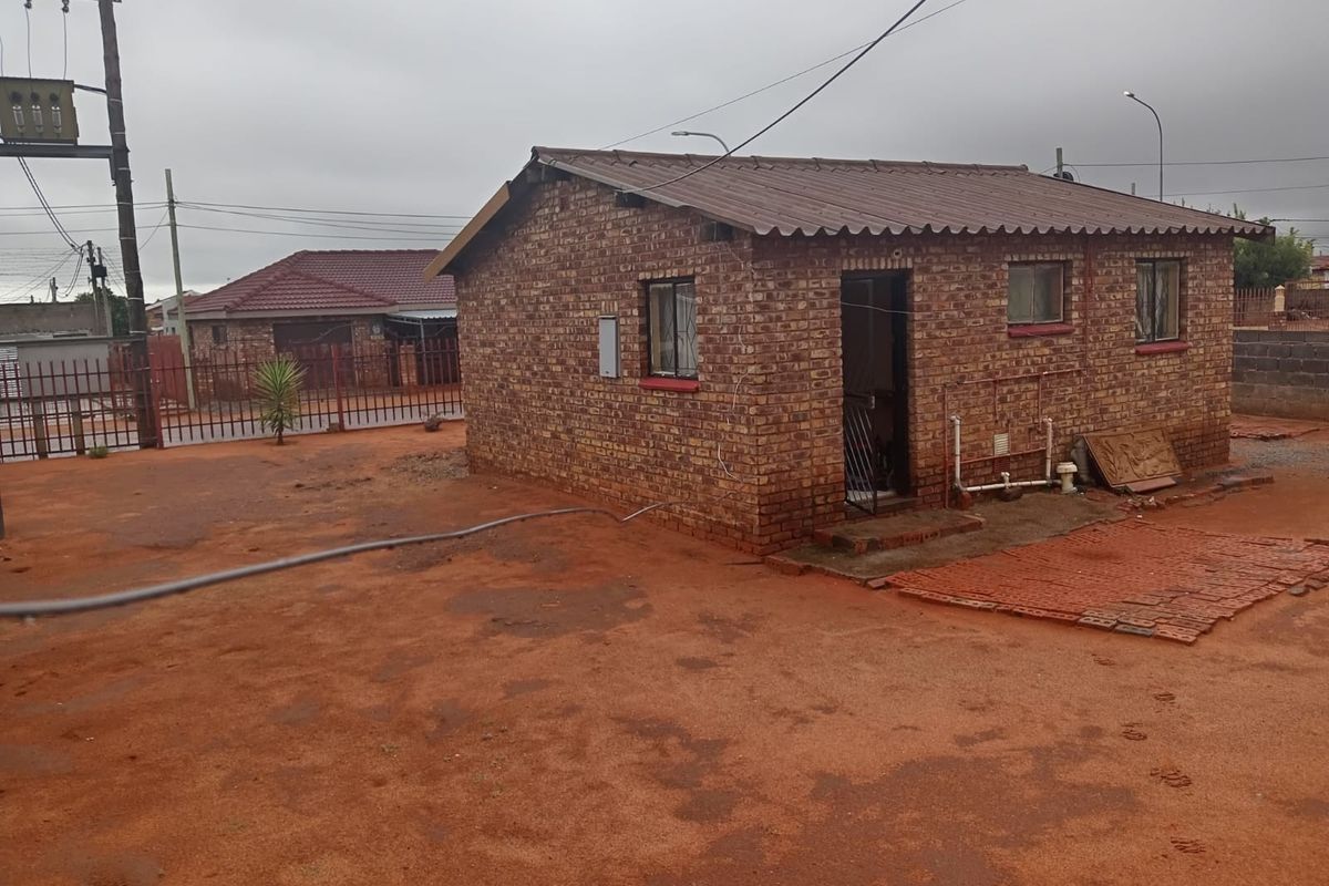 A detached face brick house featuring a corrugated metal roof and basic external infrastructure, situated on an unpaved plot with perimeter fencing.