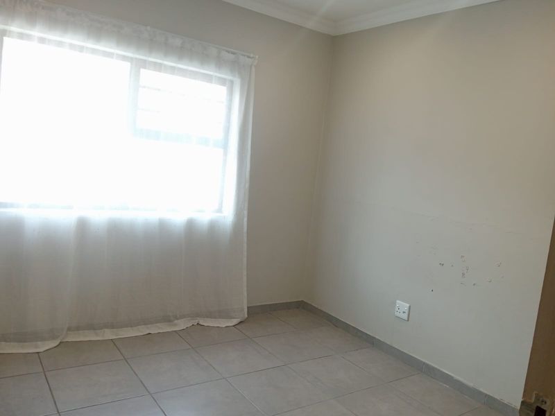 An empty bedroom featuring neutral tiled flooring, light-colored walls with visible surface wear, and a window with sheer curtains.