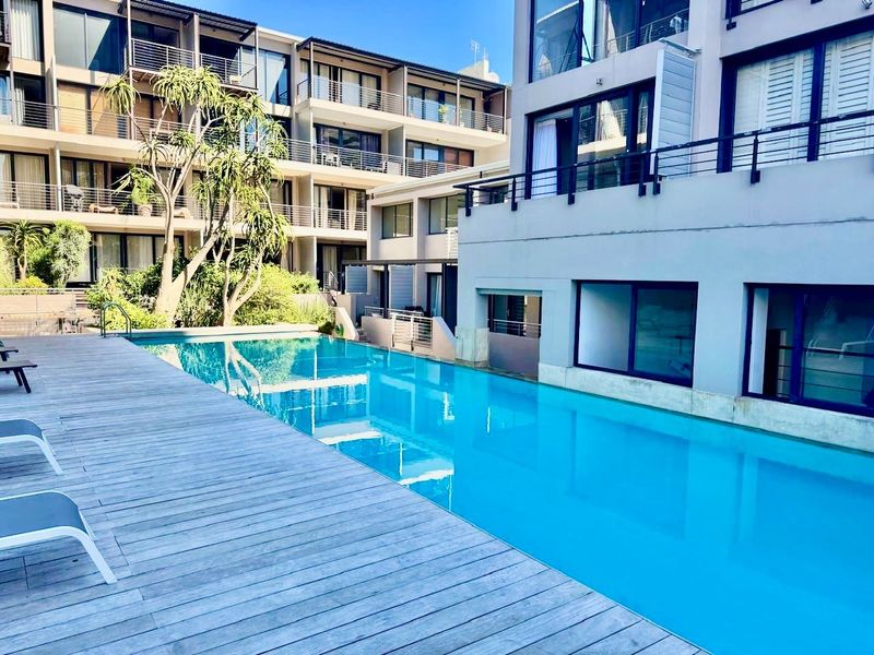 A well-maintained communal swimming pool area featuring timber decking, surrounded by modern apartment buildings with private balconies.