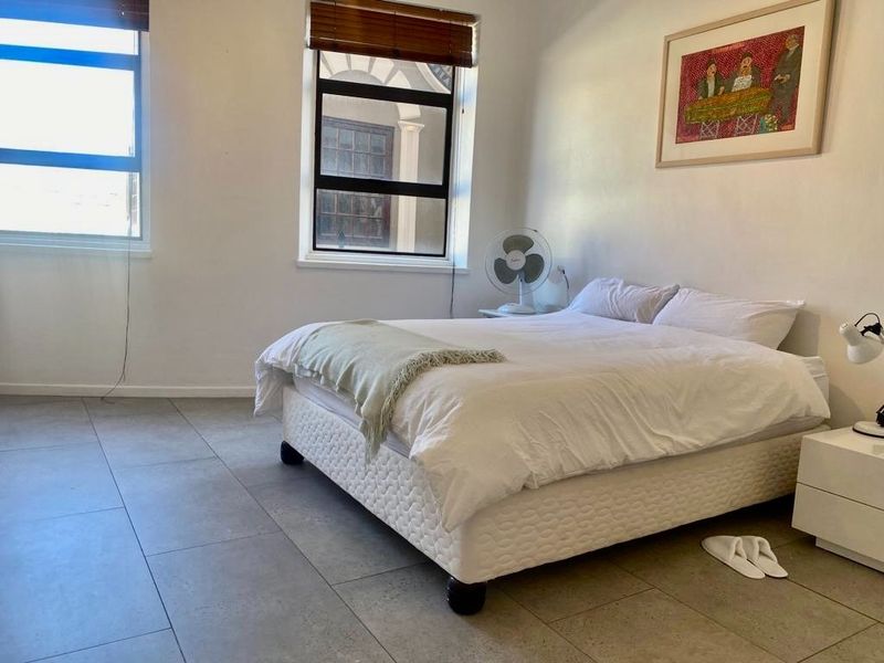 A bright bedroom featuring large windows with venetian blinds and neutral tiled flooring, offering a clean and functional living space.