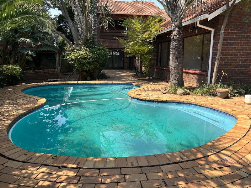 A backyard swimming pool area featuring a curved brick-paved surround, set against a face brick house with a tiled roof.