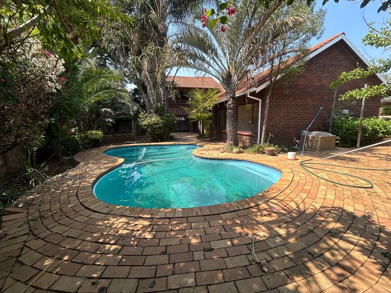 A backyard pool area featuring a kidney-shaped swimming pool surrounded by brick paving and mature garden landscaping with a face brick house exterior.