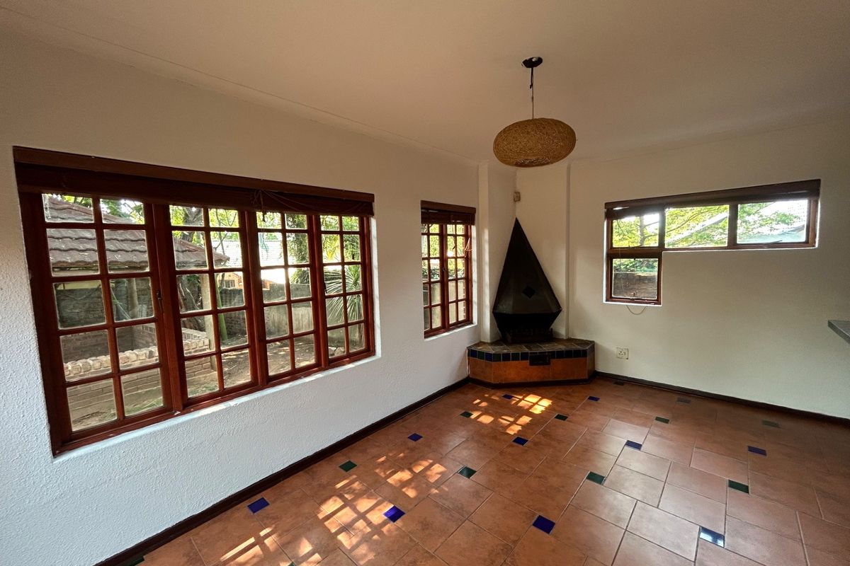 A bright, empty living room featuring terracotta tiled flooring with decorative inlays, timber-framed windows, and a unique corner fireplace.