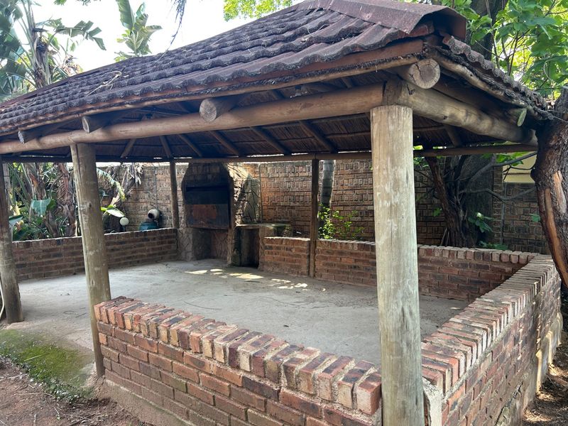 Outdoor entertainment area featuring a thatched roof, timber support structure, and a built-in brick braai facility in a garden setting.