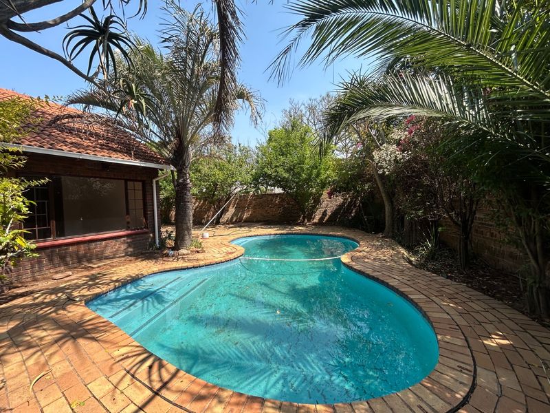 A private backyard featuring a kidney-shaped swimming pool with brick coping, surrounded by established trees and a brick boundary wall.