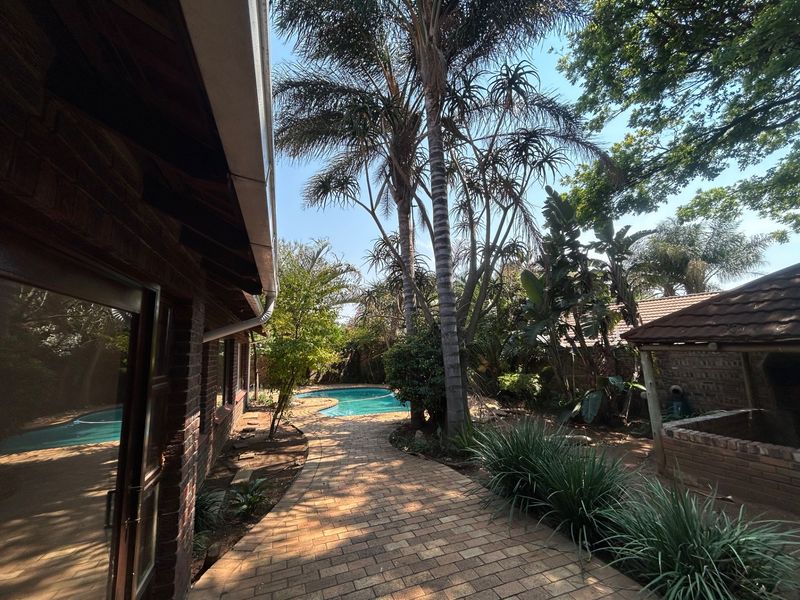 A backyard area featuring a swimming pool, brick paved walkway, and a small covered outdoor structure surrounded by mature trees and landscaping.