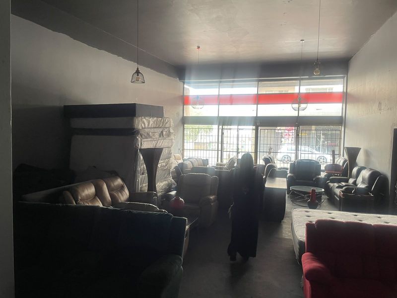 Open-plan commercial retail space featuring high ceilings, large storefront windows with security gates, and pendant lighting, suitable for a showroom or retail outlet.