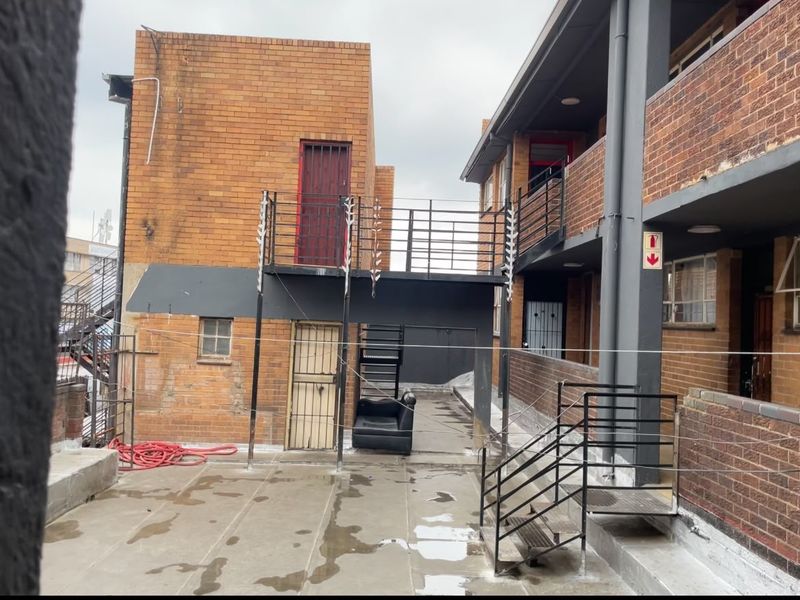 Rear exterior courtyard of a commercial building featuring brick facades, metal fire escape stairs, and security gates with paved ground surface.