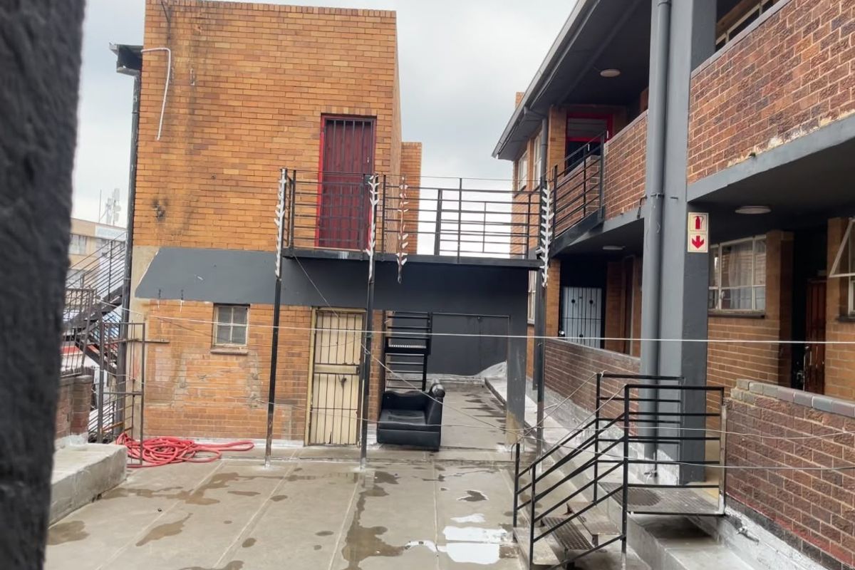 Rear exterior courtyard of a commercial building featuring brick facades, metal fire escape stairs, and security gates with paved ground surface.