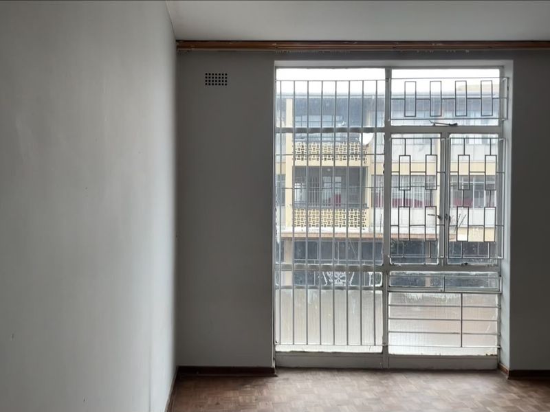 An empty room featuring parquet flooring and large windows fitted with security bars, offering a view of the adjacent building.