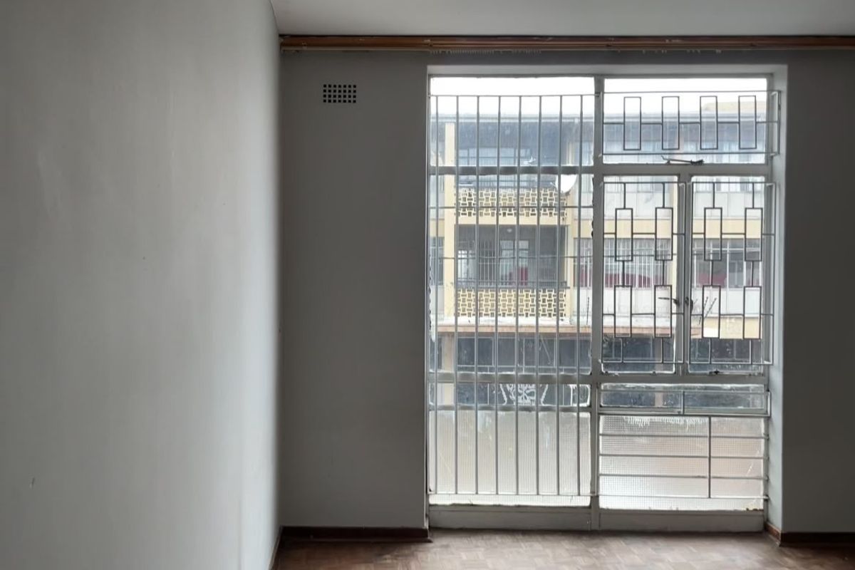 An empty room featuring parquet flooring and large windows fitted with security bars, offering a view of the adjacent building.