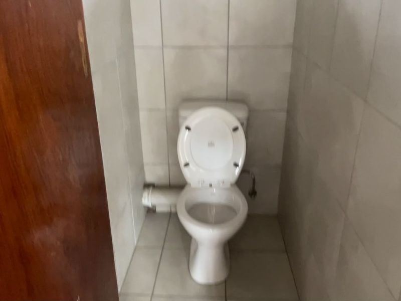 A small, functional bathroom featuring a standard toilet and tiled surfaces, currently showing signs of dated finishes and exposed plumbing.