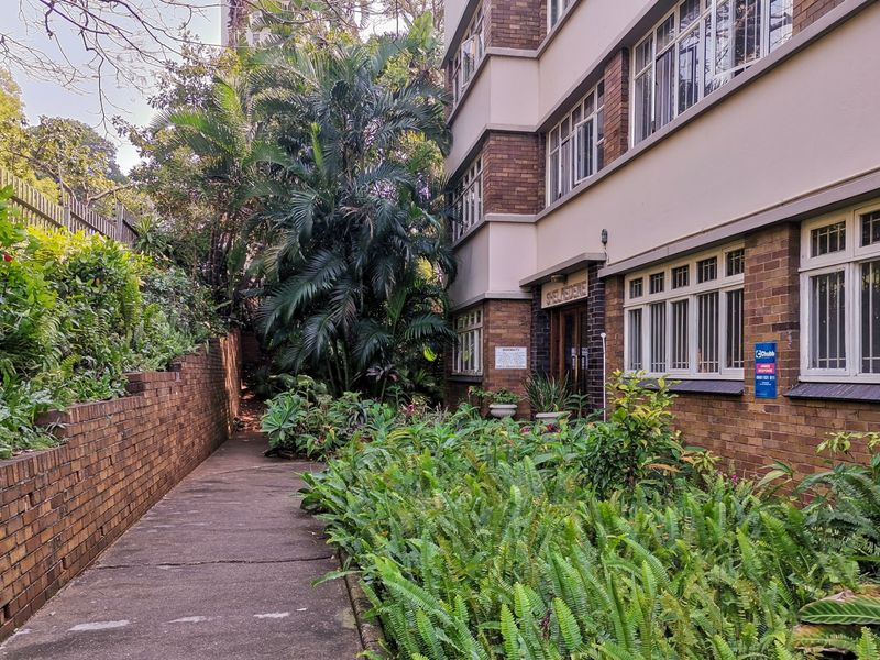 Exterior view of the apartment building entrance featuring a brick facade, established garden landscaping, and a concrete walkway leading to the main building access.