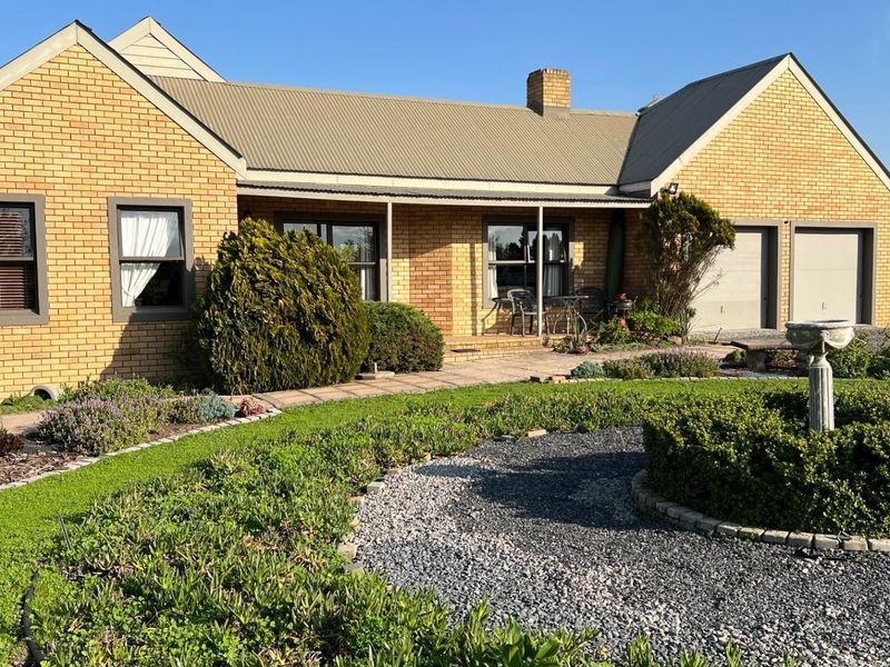A well-maintained single-story brick home featuring a metal roof, attached double garage, and a landscaped rear garden with a paved patio area.