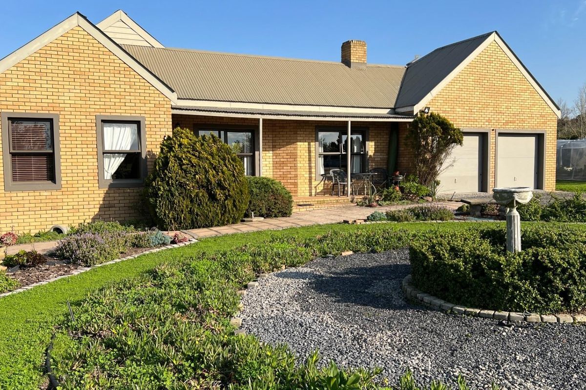 A well-maintained single-story brick home featuring a metal roof, attached double garage, and a landscaped rear garden with a paved patio area.