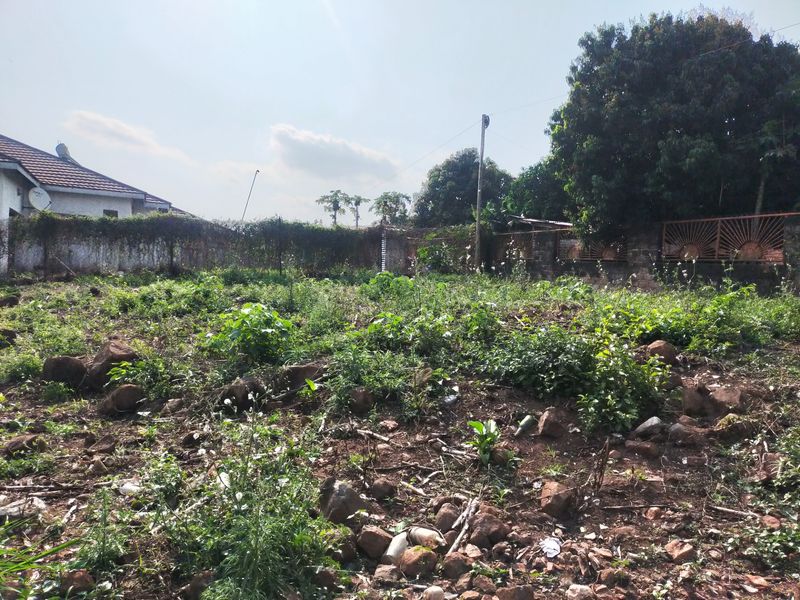 A vacant residential plot featuring overgrown vegetation, rocky terrain, and existing perimeter walling with metal fencing.