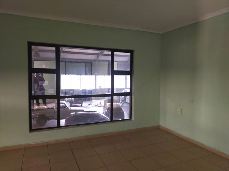 An empty office space featuring ceramic floor tiles, a large aluminum-framed window overlooking an industrial area, and light green painted walls.