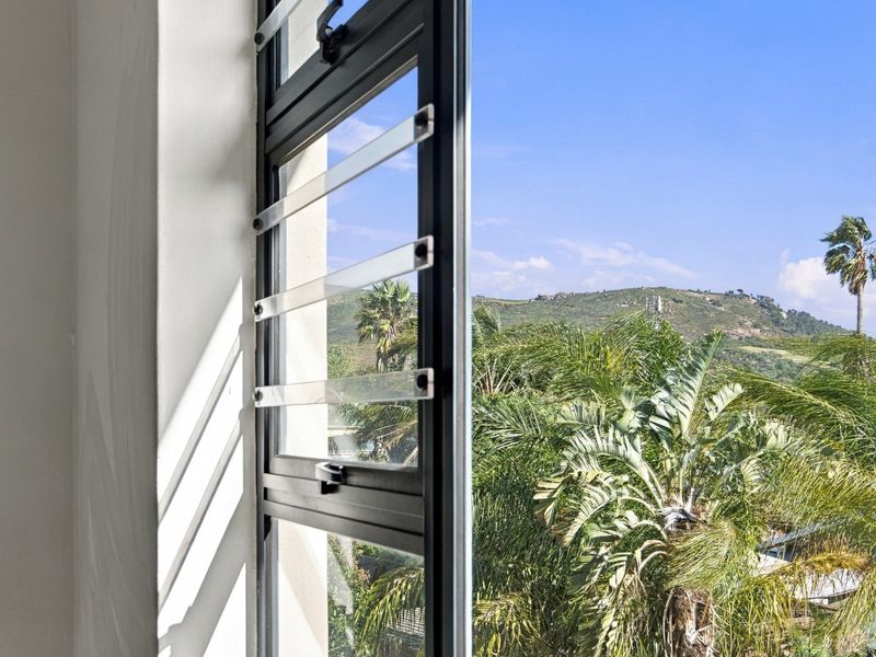 A view through a louvre window showcasing lush tropical greenery and a scenic hillside landscape.