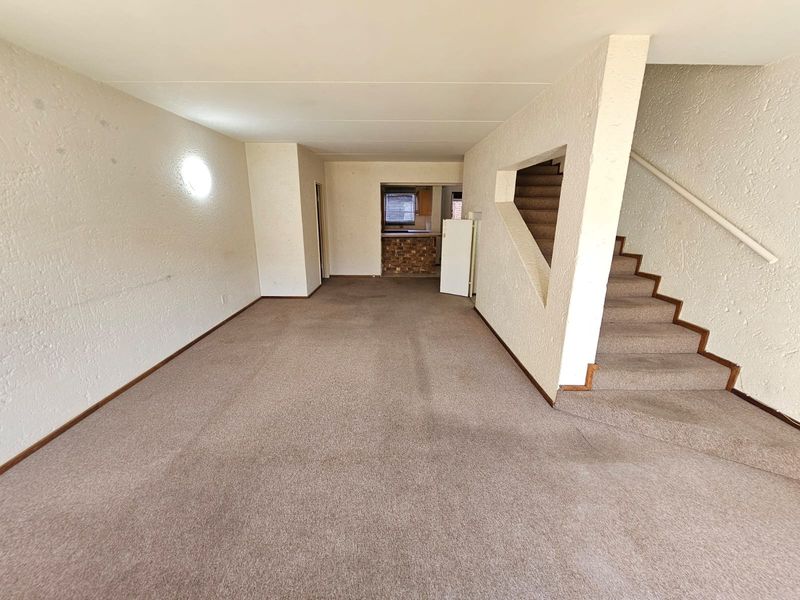 An open-plan living area featuring carpeted flooring and a staircase with a handrail, leading towards a kitchen area.