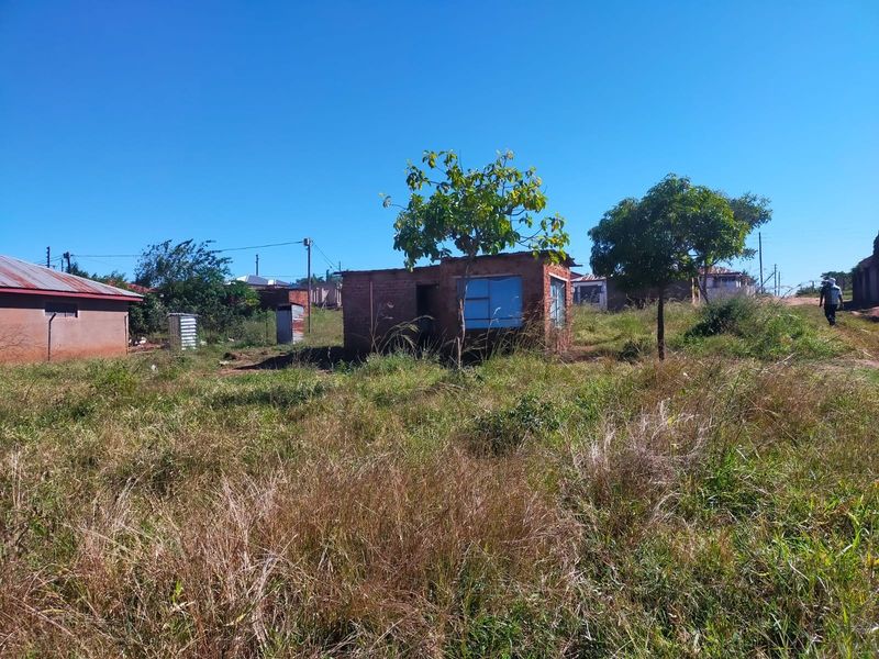 A view of a vacant residential plot featuring an unfinished small brick structure amidst overgrown grass and mature trees.