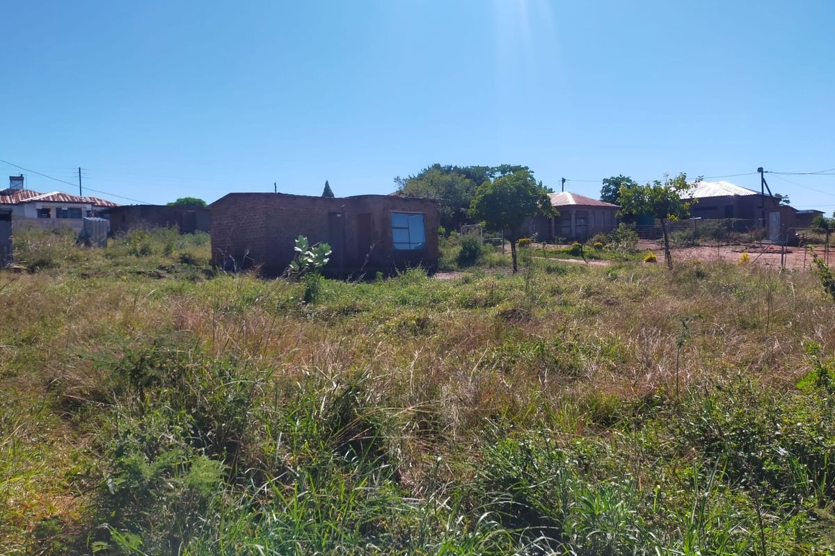 A view of a residential plot featuring an existing small brick structure surrounded by overgrown vegetation and open land.