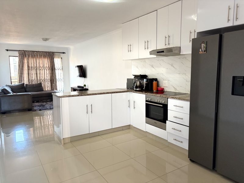 Modern open-plan kitchen featuring white cabinetry, granite countertops, and integrated appliances, seamlessly flowing into the adjacent living area with tiled flooring throughout.