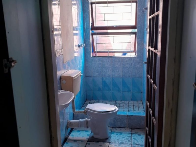 A compact bathroom featuring blue tiled walls and flooring, a standard toilet, washbasin, and a shower area with security bars on the window.