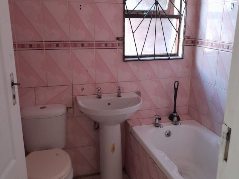 A dated bathroom featuring pink wall tiles, a pedestal basin, a standard toilet, and a bathtub with security bars on the window.