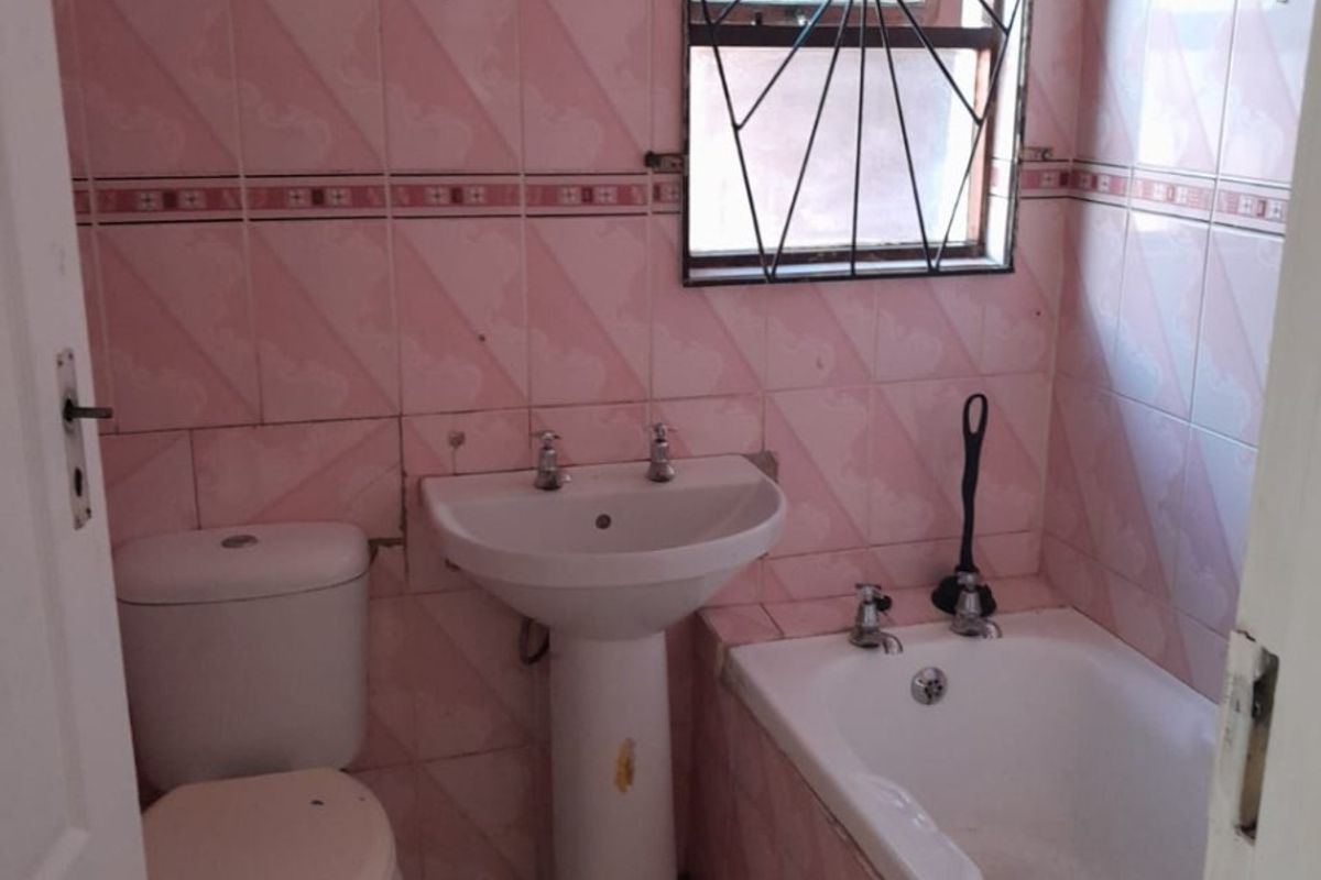 A dated bathroom featuring pink wall tiles, a pedestal basin, a standard toilet, and a bathtub with security bars on the window.