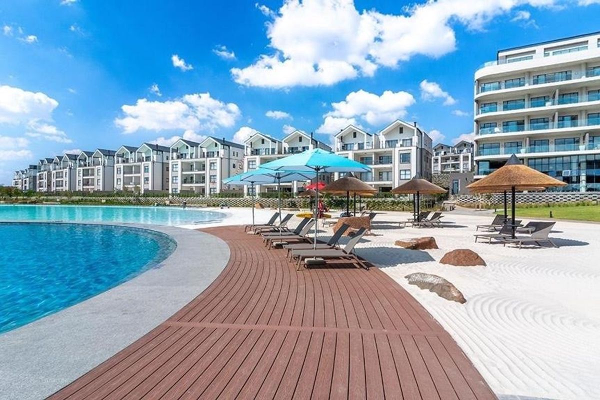 A well-maintained communal pool area featuring a swimming pool, timber decking, and a sandy beach section within a modern apartment complex.