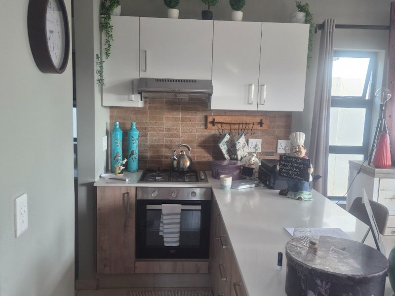 A compact, modern kitchen featuring light wood-finish cabinetry, stone countertops, a built-in oven with gas hob, and a decorative tiled backsplash.