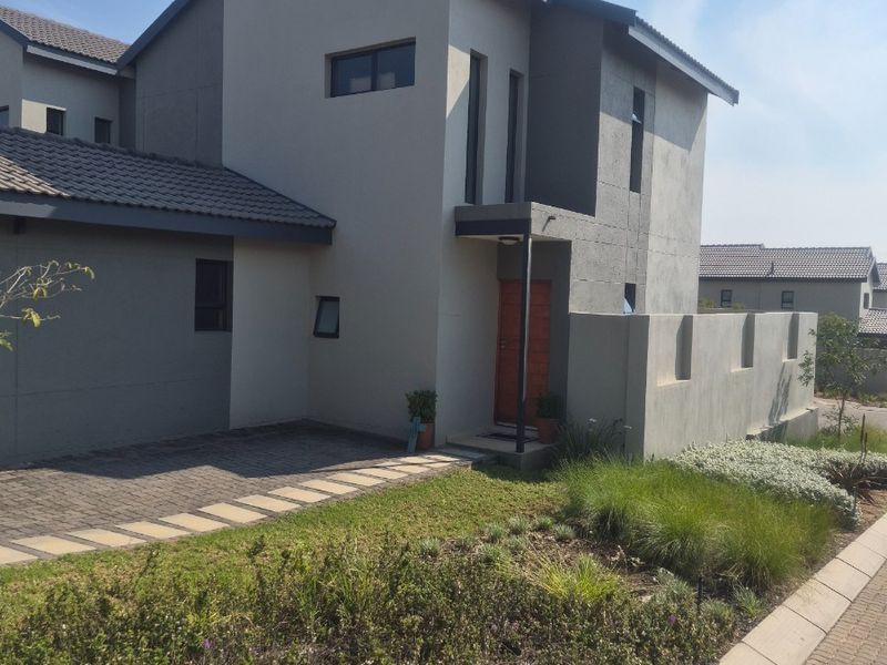 A modern double-storey townhouse featuring a paved driveway, landscaped garden, and a contemporary facade with a timber entrance door.
