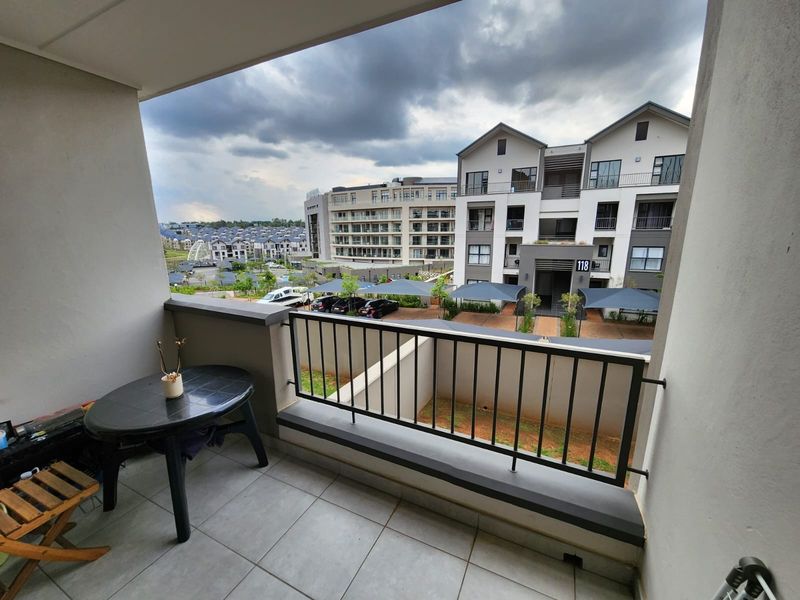 A covered balcony featuring tiled flooring and a metal balustrade, offering an elevated view of the surrounding apartment complex and parking area.