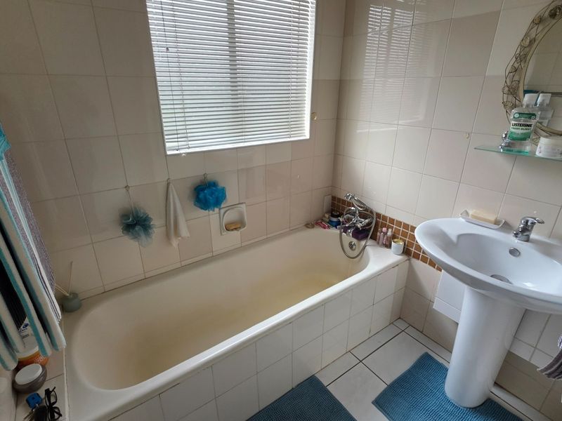A functional bathroom featuring a pedestal basin, bathtub with mixer tap, and neutral wall tiling with a window for natural light.