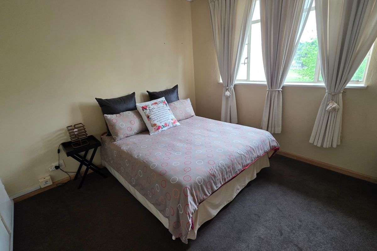 A well-lit bedroom featuring neutral wall tones, carpeted flooring, and large windows with curtains, providing a comfortable and functional living space.