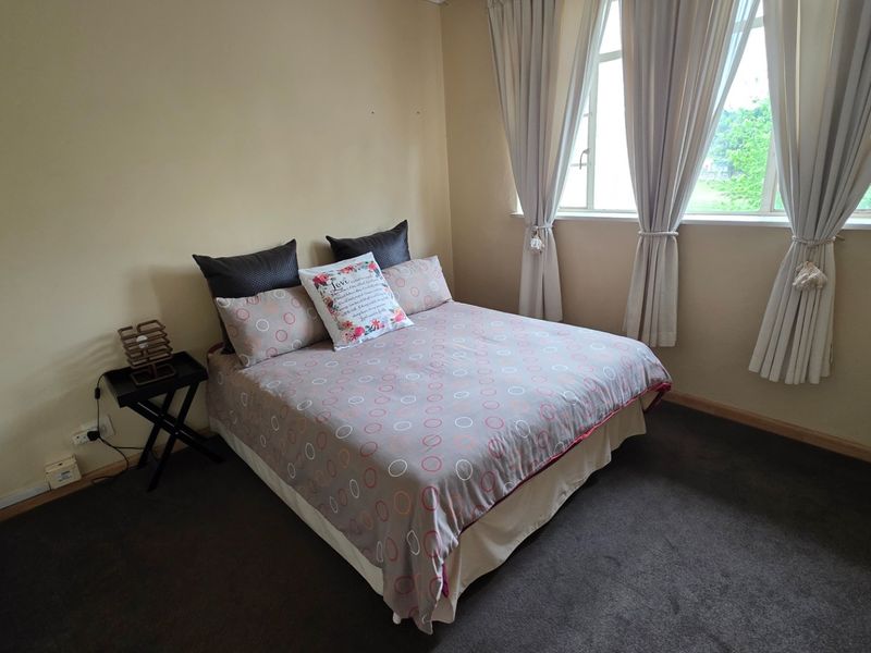 A well-lit bedroom featuring neutral wall tones, carpeted flooring, and large windows with curtains, providing a comfortable and functional living space.