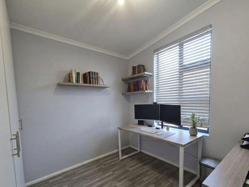 A neat home office featuring laminate flooring, built-in cupboard storage, and venetian blinds, providing a functional workspace with good natural light.