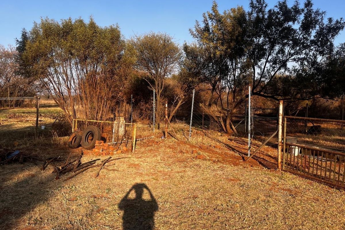A view of a residential plot featuring mature trees, wire fencing, and a metal gate, showing signs of neglect and overgrowth.