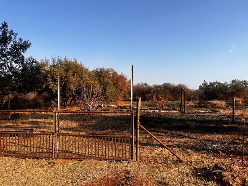A view of a vacant residential plot featuring a weathered metal gate and perimeter wire fencing with natural vegetation.