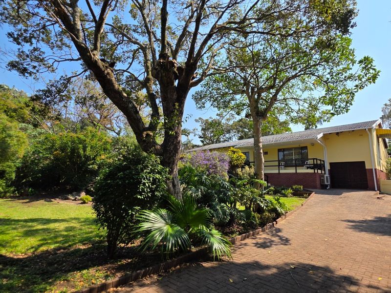Front view of the property featuring a paved driveway, established garden, and a single-story house with a tiled roof and brick base.