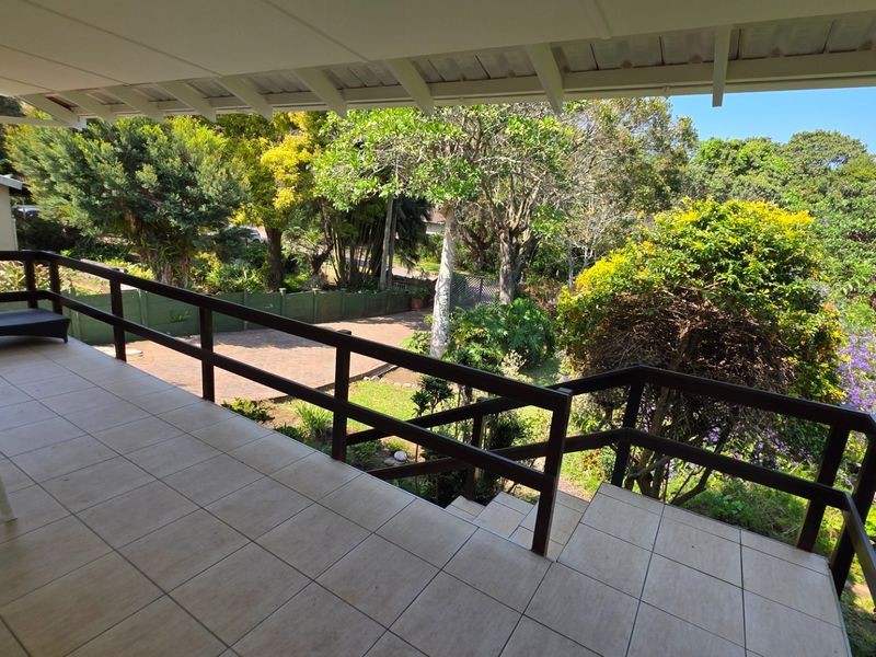 A spacious covered balcony featuring tiled flooring and a timber balustrade, overlooking a lush garden area.